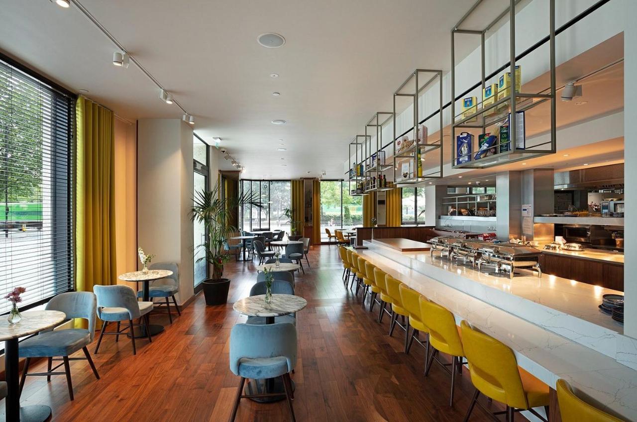 Tower Suites By Blue Orchid Londra Exterior foto The photo shows a modern restaurant interior. It features a spacious layout with large windows allowing natural light to flood the space. The decor includes a combination of wooden flooring and colorful furniture—blue and yellow chairs arranged aroun