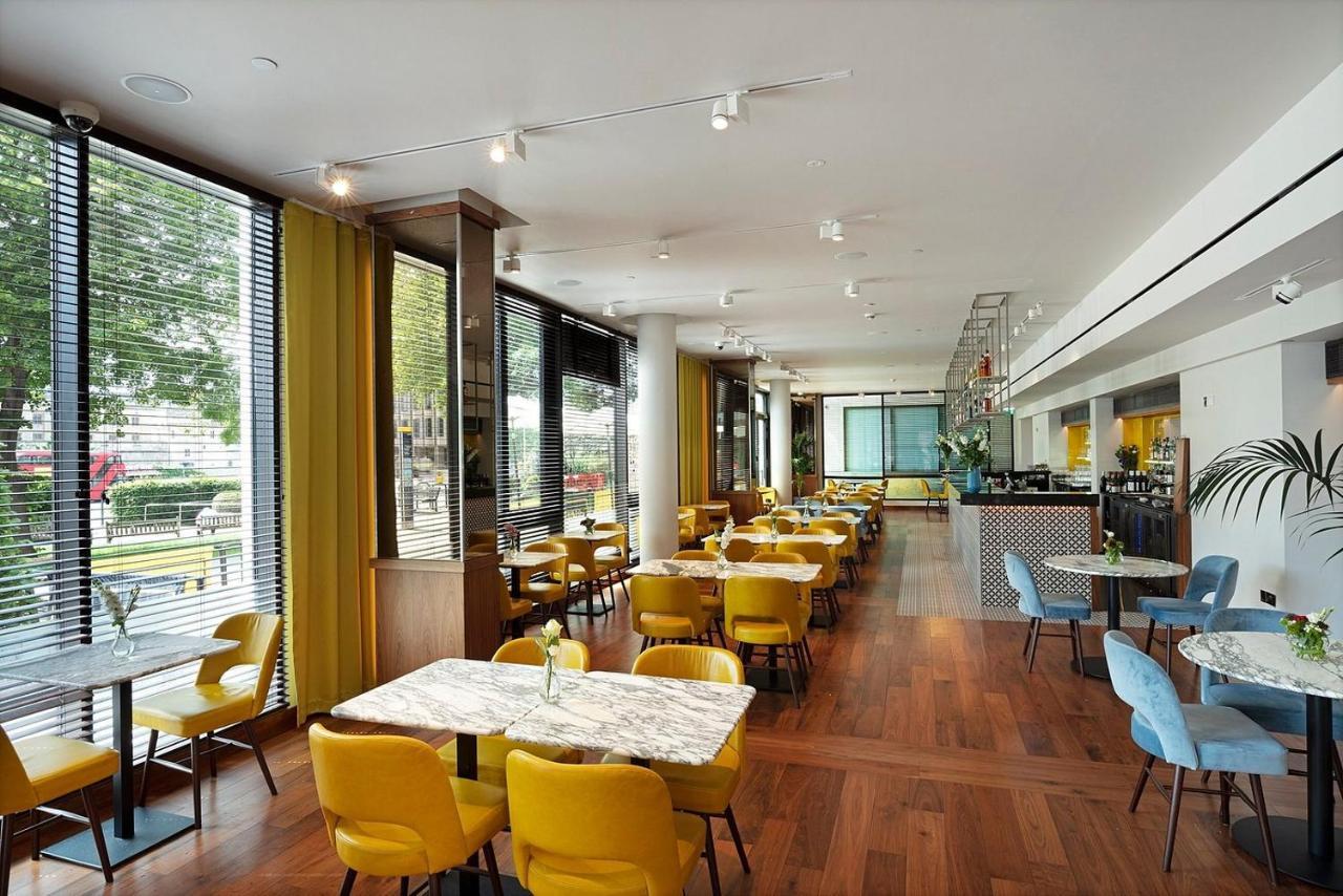 Tower Suites By Blue Orchid Londra Exterior foto The photo depicts a modern dining area with a bright and airy atmosphere. There are several tables with marble tops, accompanied by yellow chairs, creating a vibrant contrast with the wood flooring. The space features large windows that allow ample n