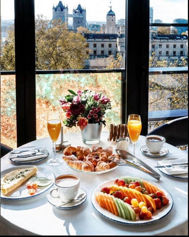 Tower Suites By Blue Orchid Londra Exterior foto The photo showcases a beautifully set breakfast table with a scenic view through large glass windows. The table features an array of items, including:

1. A small vase filled with colorful flowers.
2. Two glasses of orange juice.
3. A cup of coffee o