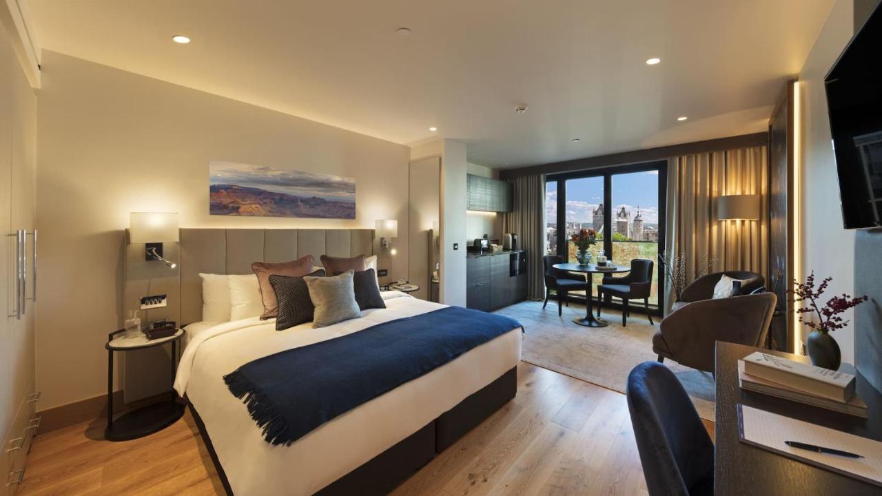 Tower Suites By Blue Orchid Londra Exterior foto The photo shows a modern hotel room. It features a large bed with a dark blue throw blanket and several decorative pillows. The walls are painted in neutral tones, and there's a landscape painting above the bed. To one side, there is a small kitchene