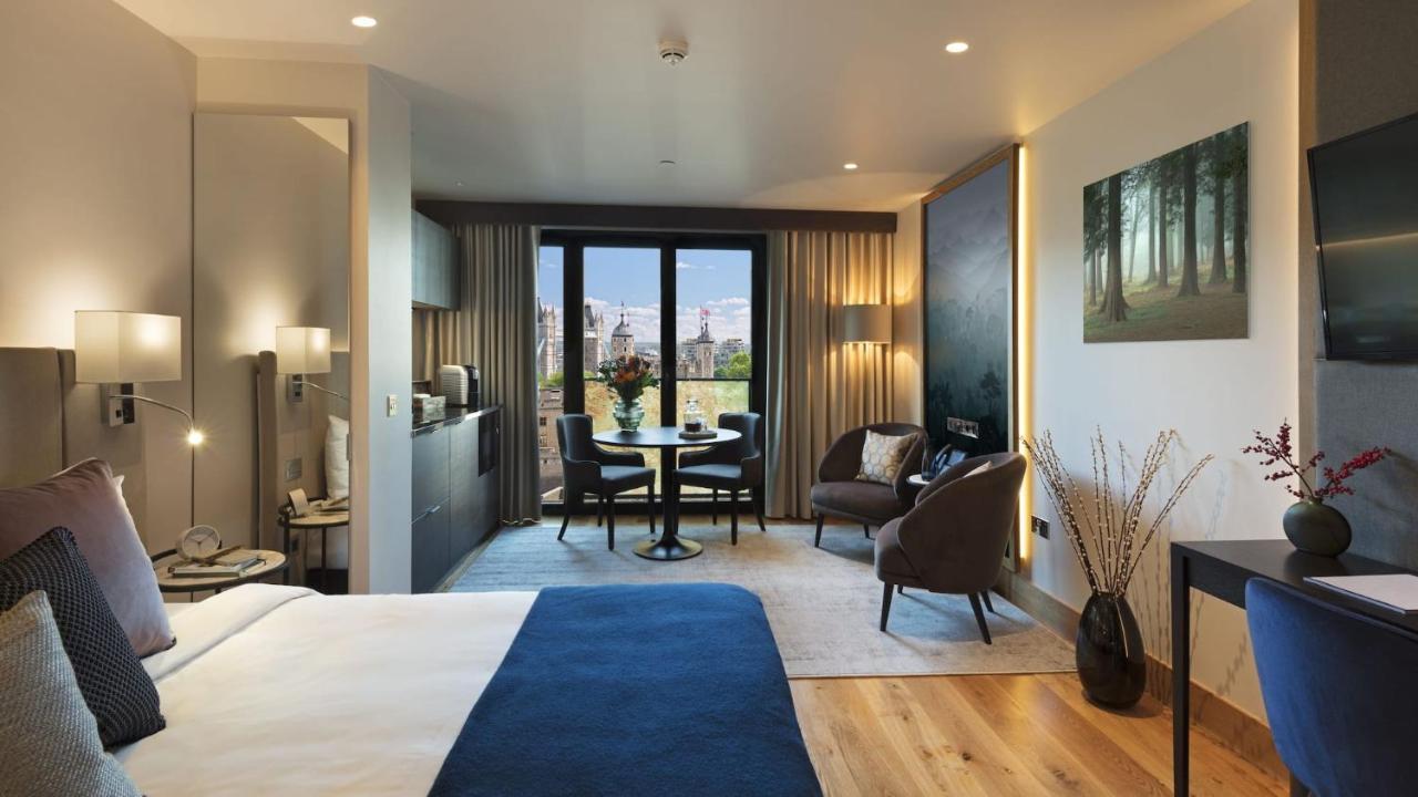 Tower Suites By Blue Orchid Londra Exterior foto The photo shows a modern hotel room designed with a contemporary aesthetic. On one side, there is a large bed with a blue blanket, while the other side features a sitting area with two chairs and a small round dining table. Large windows let in natur