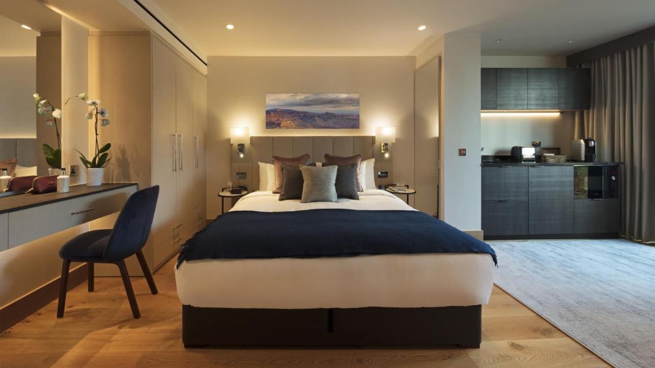Tower Suites By Blue Orchid Londra Exterior foto The photo displays a modern hotel room featuring a plush bed with a dark blue bedspread and several decorative pillows. The walls are adorned in neutral tones, and there is a piece of artwork above the bed depicting a serene landscape. On the side, t