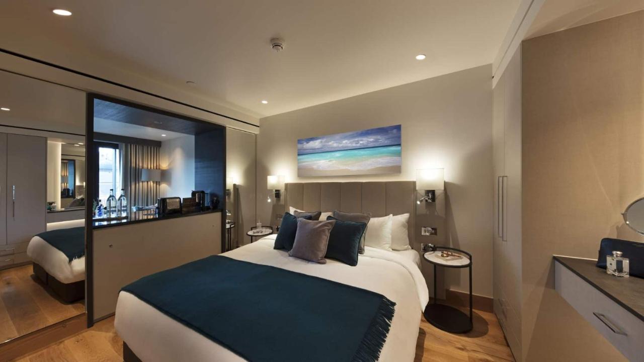 Tower Suites By Blue Orchid Londra Exterior foto The photo shows a modern hotel bedroom featuring a large bed with a light-colored headboard and a deep blue throw at the foot. The bed is adorned with multiple decorative pillows. On the wall above the bed, there is a colorful painting depicting a be