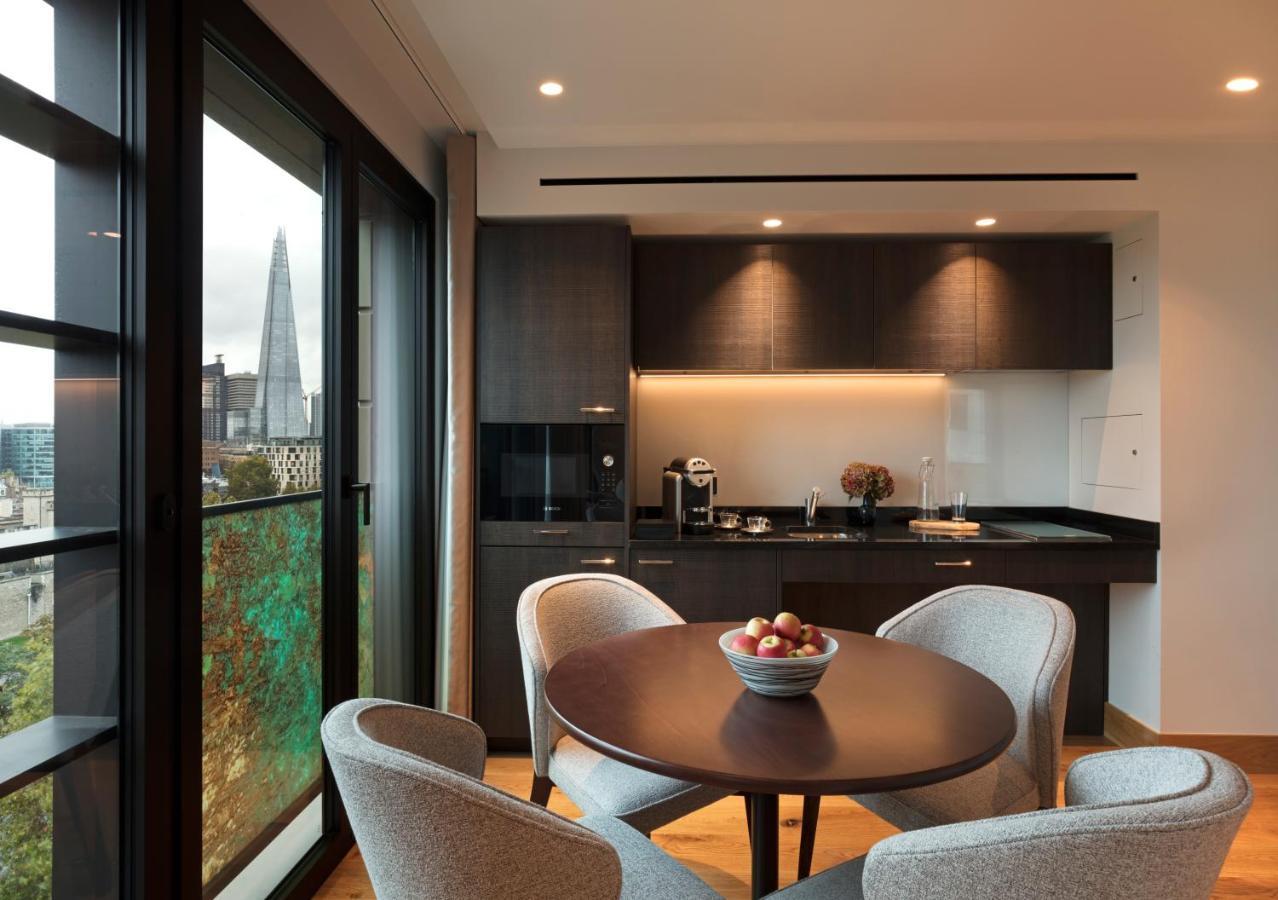 Tower Suites By Blue Orchid Londra Exterior foto The photo shows a modern kitchen and dining area. It features a round table with four gray chairs, and a bowl of apples is placed in the center of the table. The kitchen has dark cabinetry, a black countertop, and built-in appliances. Large windows a
