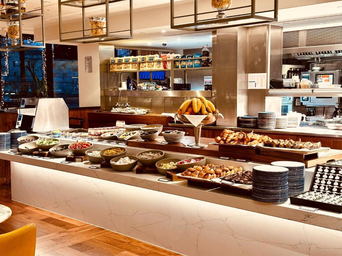Tower Suites By Blue Orchid Londra Exterior foto The photo depicts a buffet-style setup, likely in a restaurant or a hotel dining area. In the foreground, there are multiple bowls and platters filled with a variety of food items, including salads, fruits, pastries, and other dishes. There is a long