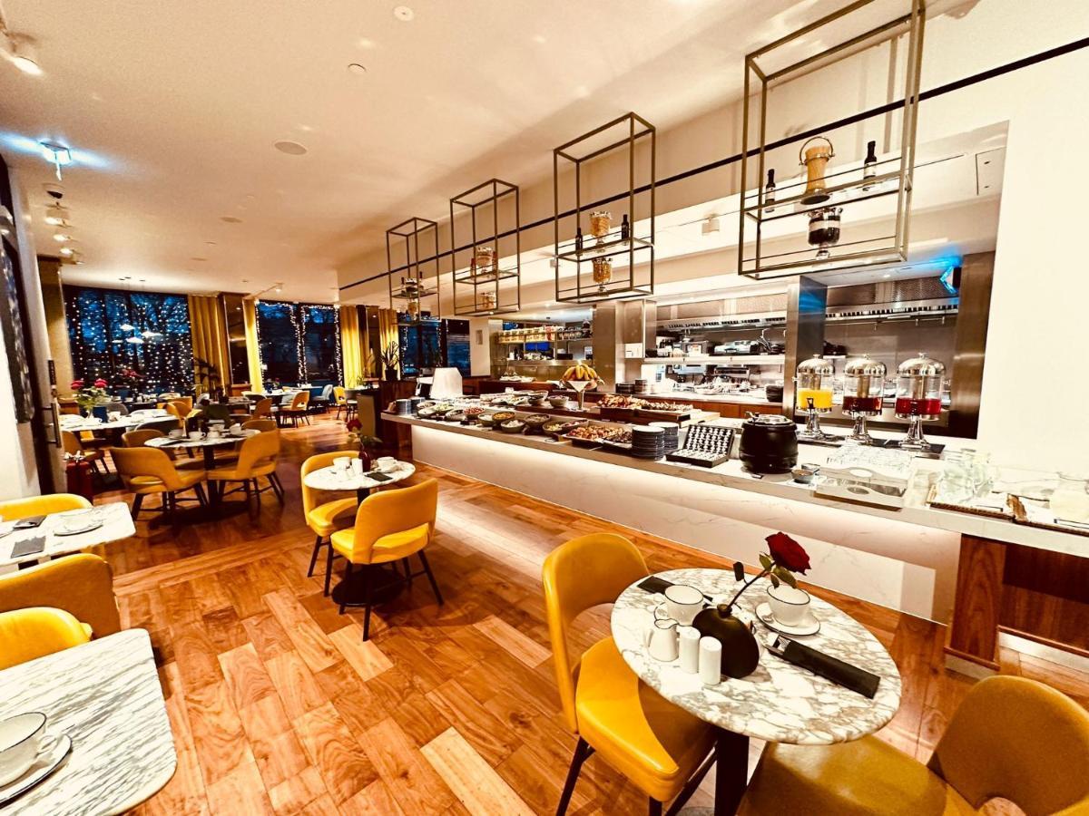 Tower Suites By Blue Orchid Londra Exterior foto The photo depicts a modern restaurant interior with a spacious layout. There are several round tables, each adorned with marble tabletops and stylish yellow chairs. A buffet area is visible in the background, featuring various food setups and kitchen