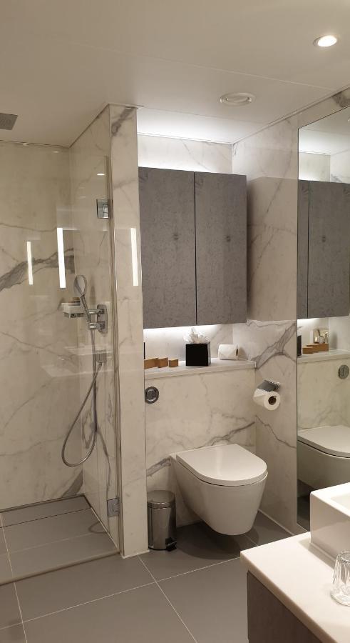 Tower Suites By Blue Orchid Londra Exterior foto The photo shows a modern bathroom design featuring elegant marble walls. On one side, there is a transparent shower enclosure with a showerhead and controls visible. Opposite the shower, there is a toilet, and above it are some sleek cabinets. The sp